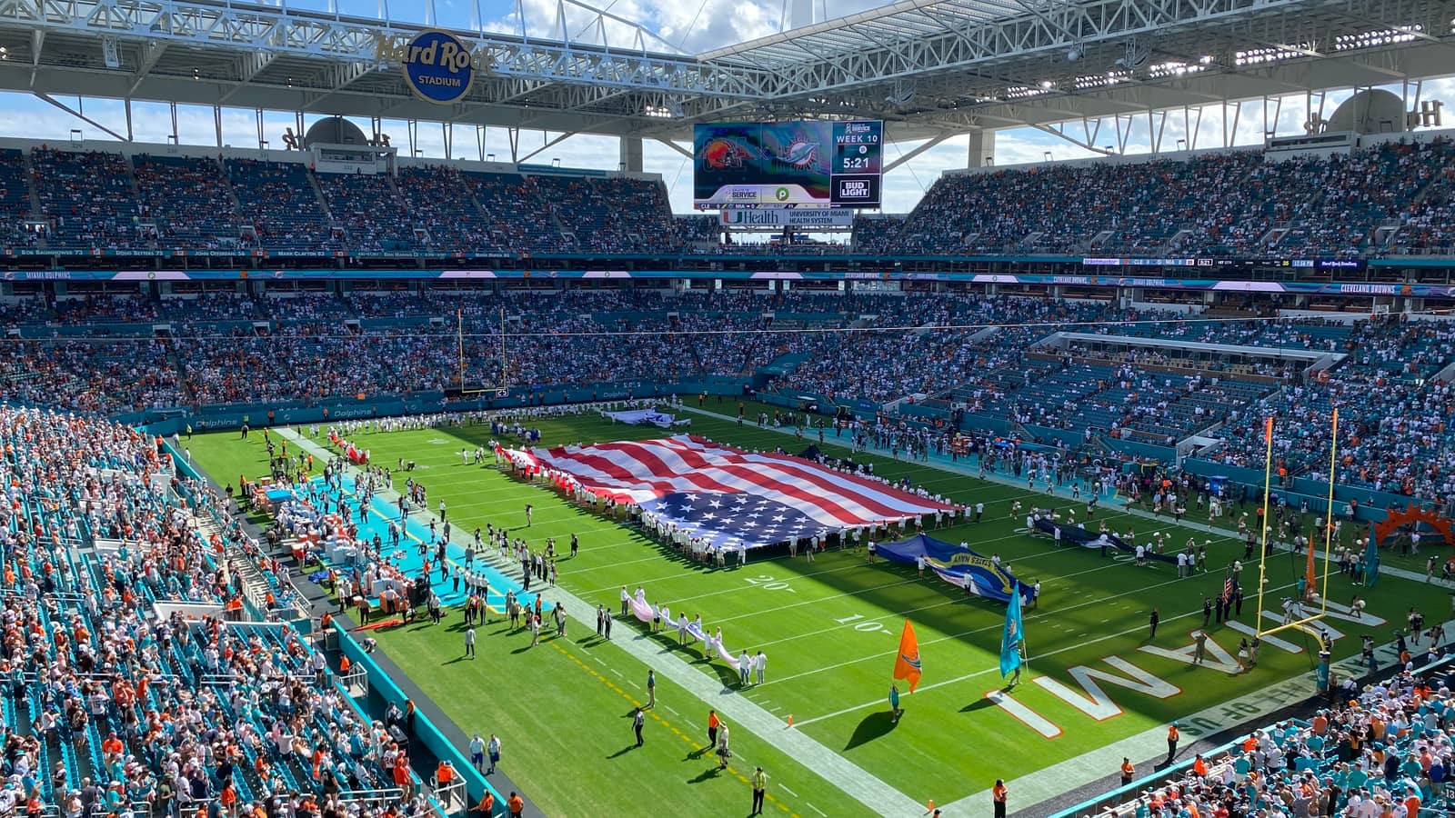 A packed Hard Rock Stadium in Miami during the national anthem before a Dolphins game, a giant American flag held across the field